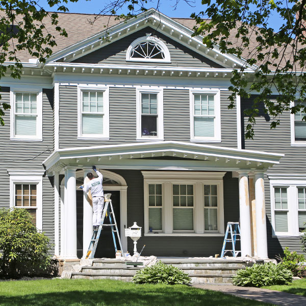 Painter on a ladder painting white trim on a gray two-story home