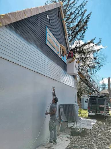 Painter spraying the siding of a house while a second painter works from a lift